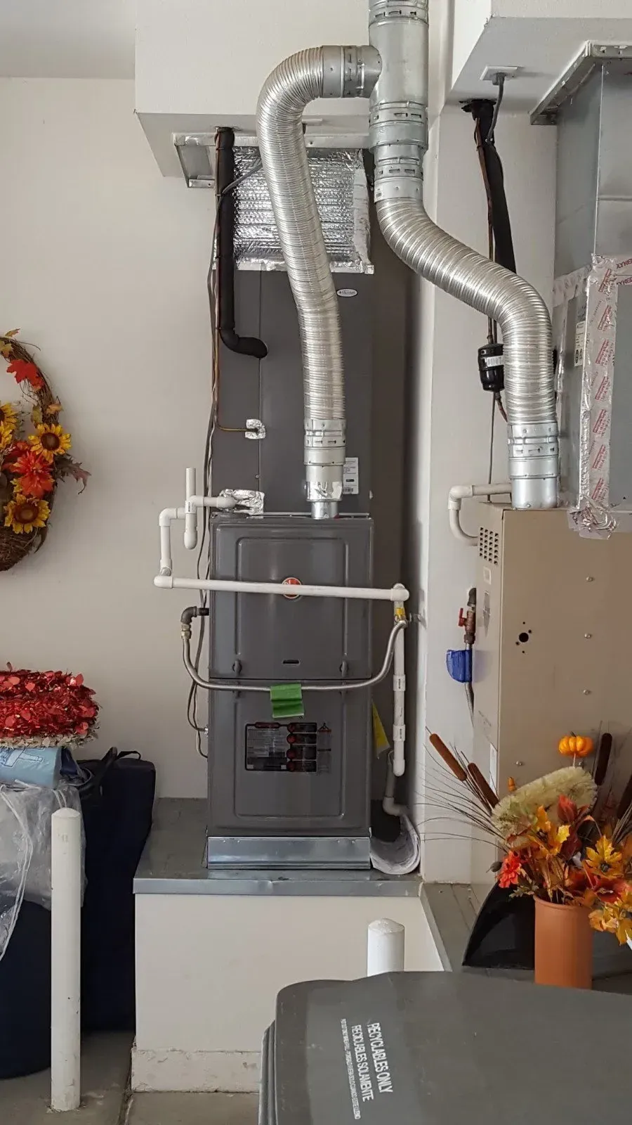 Furnace in a garage with silver ducts and pipes. Unit is grey and set on a metal platform.