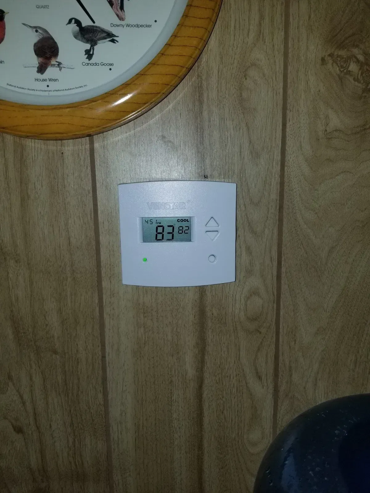 Thermostat on wood-paneled wall, displaying 83 degrees. A clock is above the thermostat.