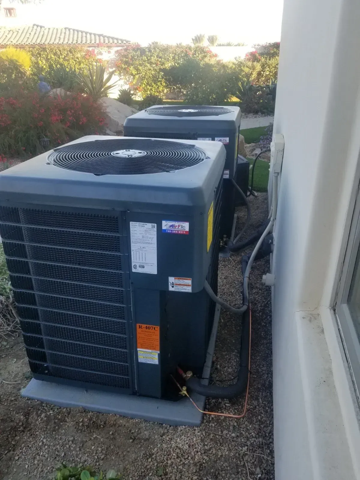 Two dark-colored air conditioning units side-by-side next to a building wall outdoors.