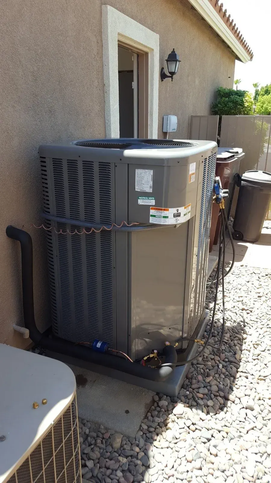 Gray air conditioning unit outside a building with a white door and trash can.