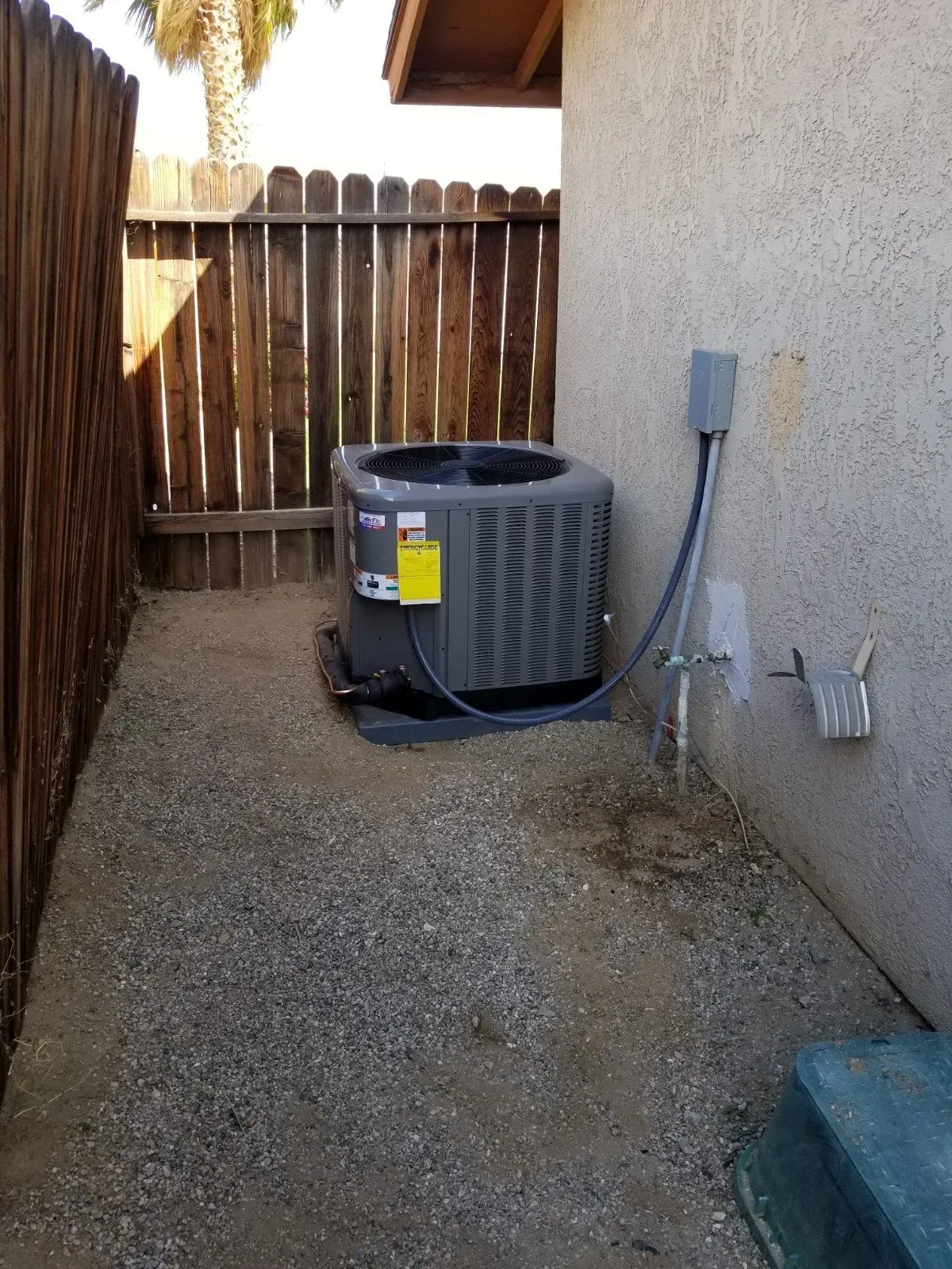 An air conditioning unit sits next to a textured beige building and wooden fence on a gravel path.