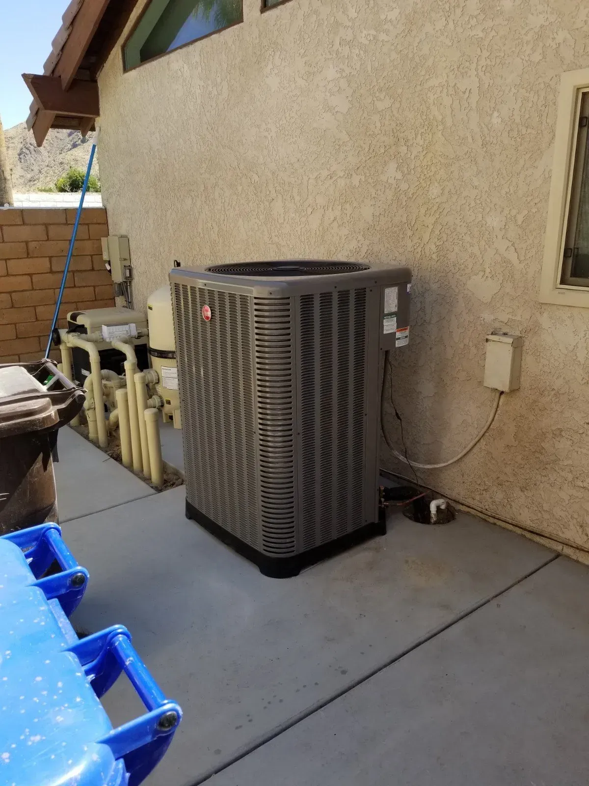 Air conditioning unit outside a stucco building, next to pool equipment.