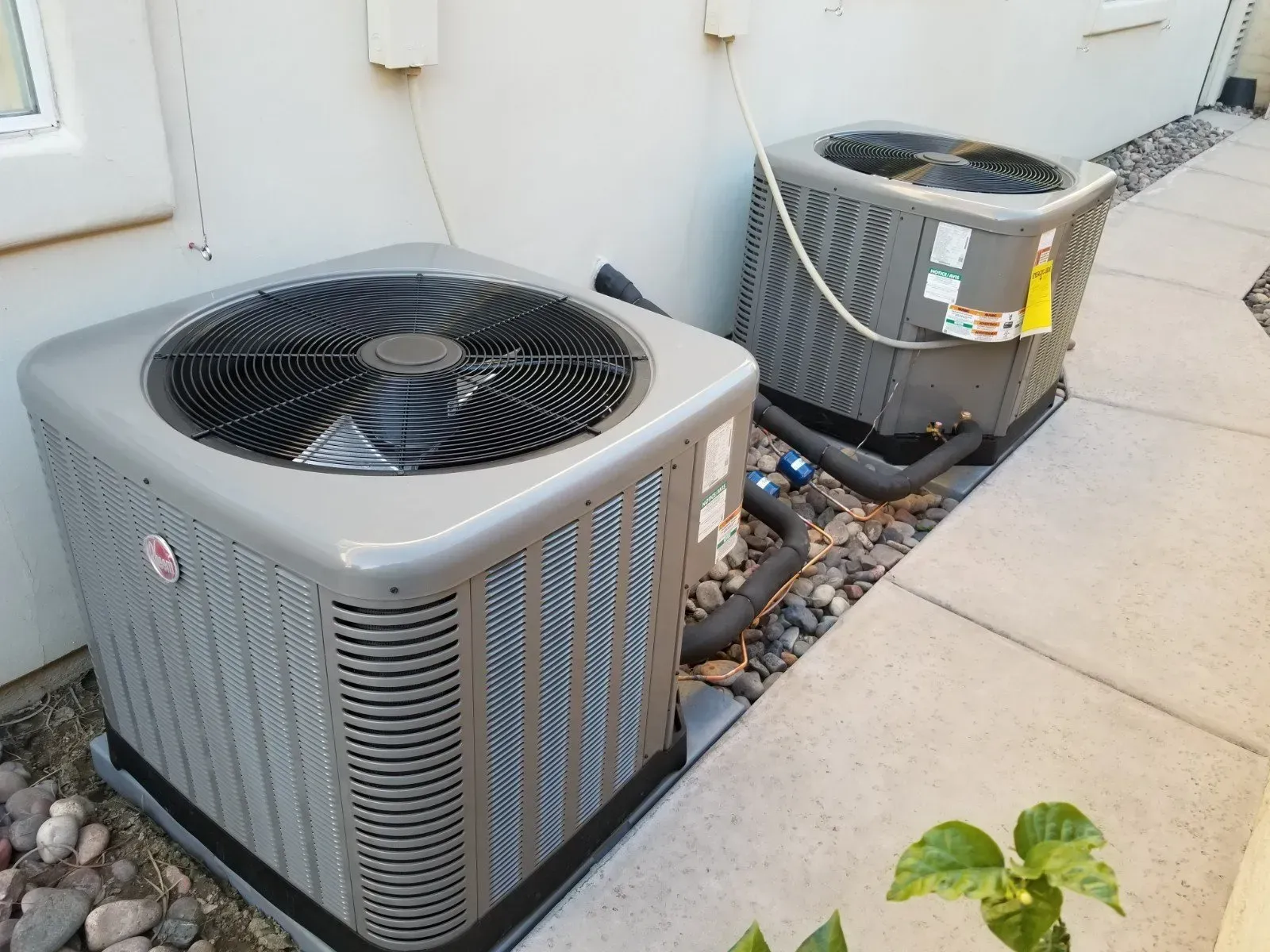 Two gray air conditioning units next to a building wall, on a concrete path with rocks.