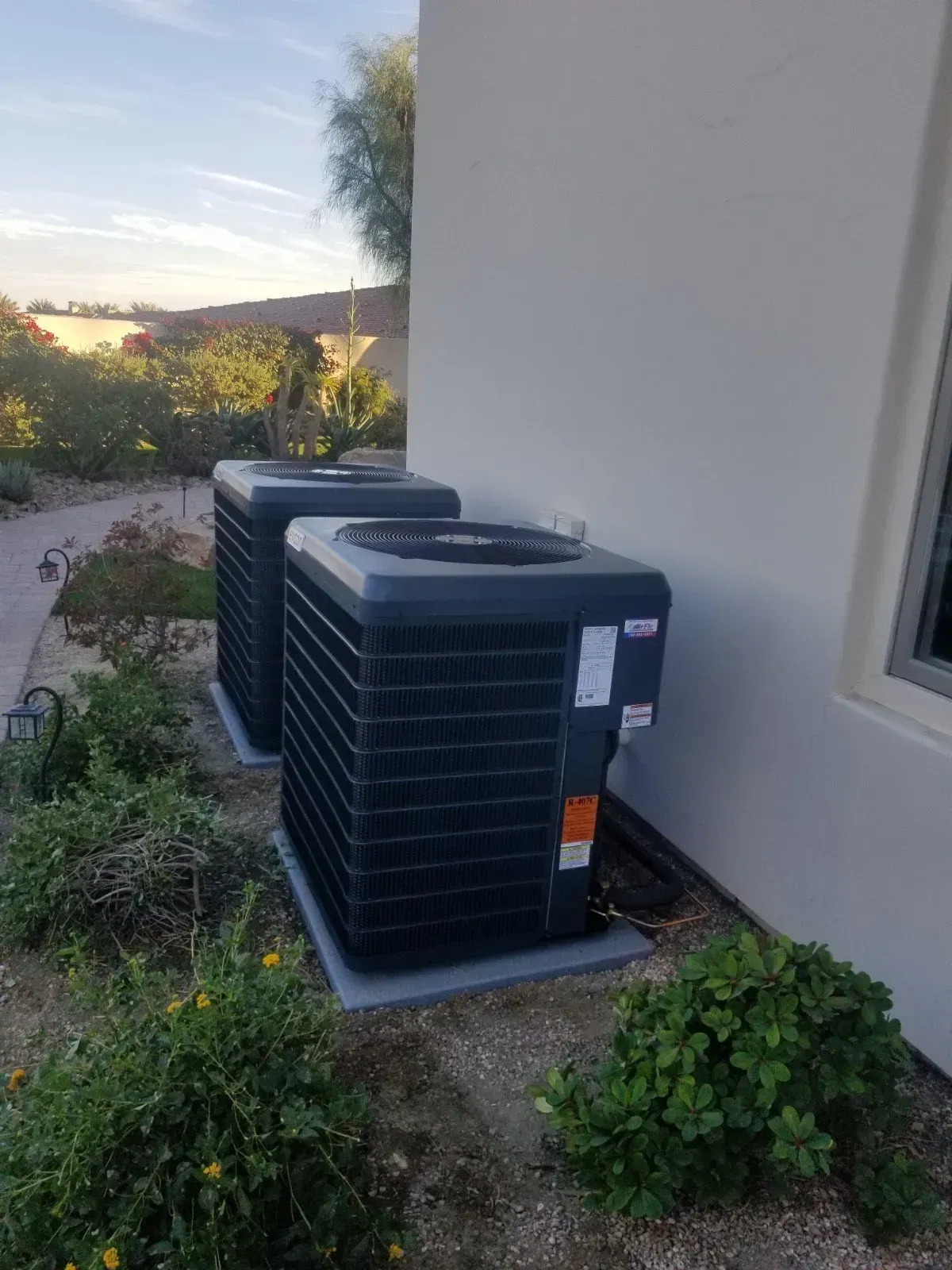 Two dark-colored air conditioning units next to a light-colored building, surrounded by plants and shrubbery.