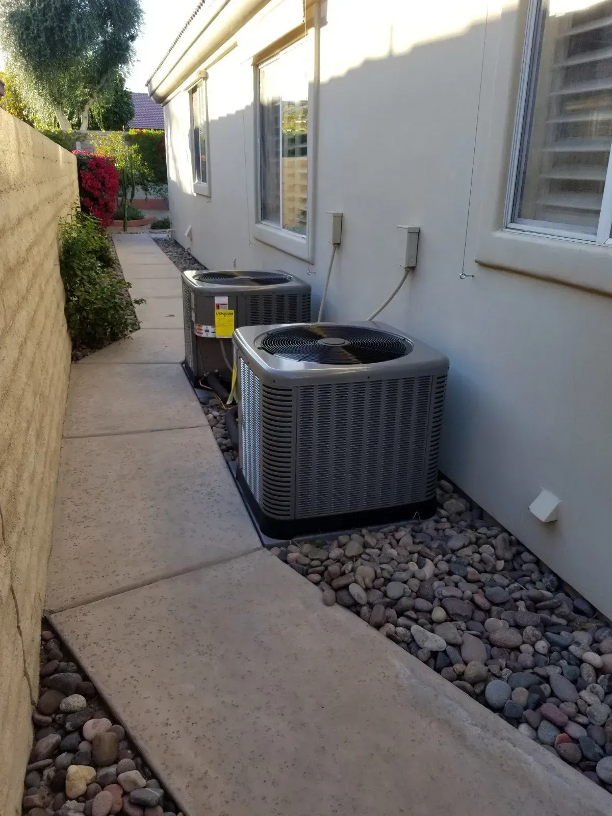 Two air conditioning units sit on a bed of rocks next to a building and concrete walkway.