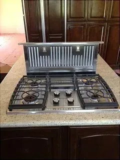 Stainless steel gas cooktop with four burners, grates, and integrated range hood in a kitchen setting.