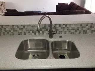 Stainless steel double sink with faucet, white countertop, and tiled backsplash.