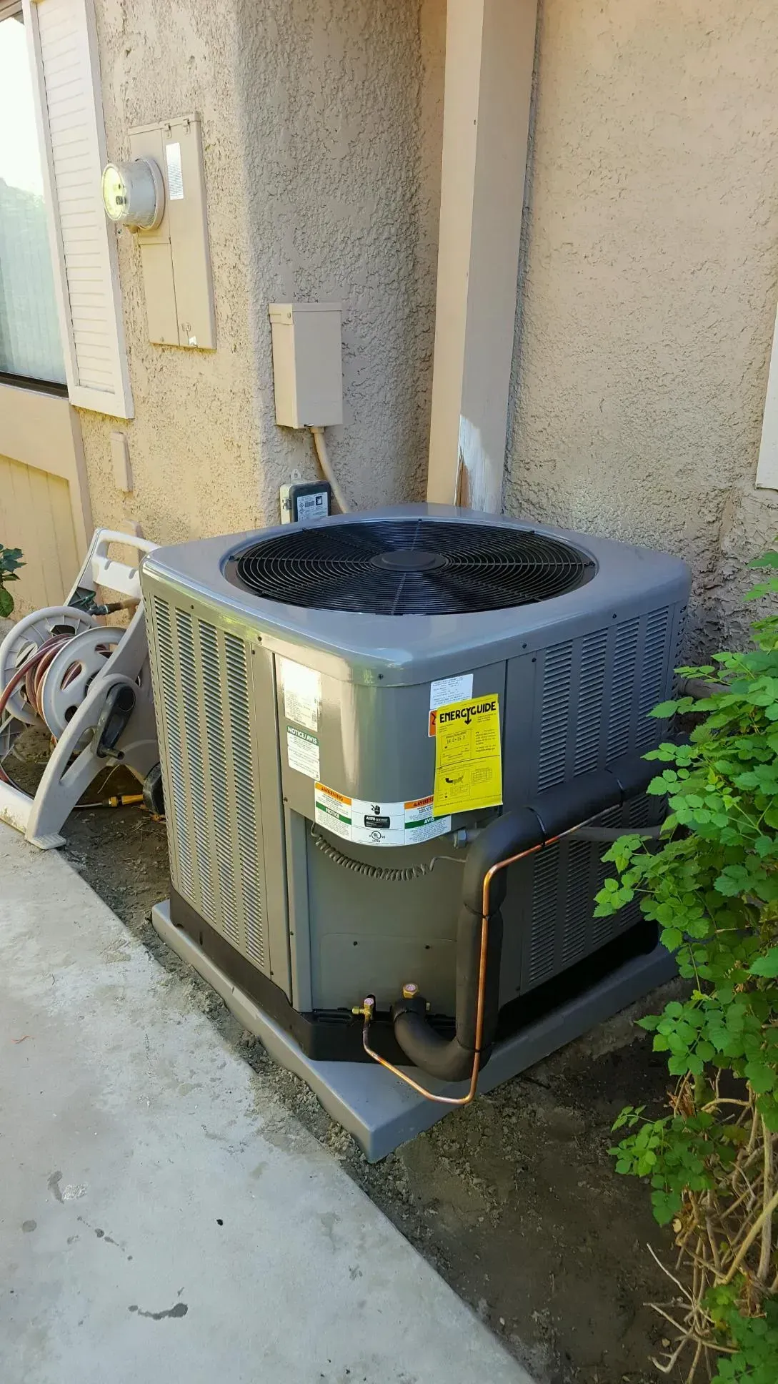 Air conditioning unit outside a building, gray with copper piping, sitting on a concrete pad next to bushes.