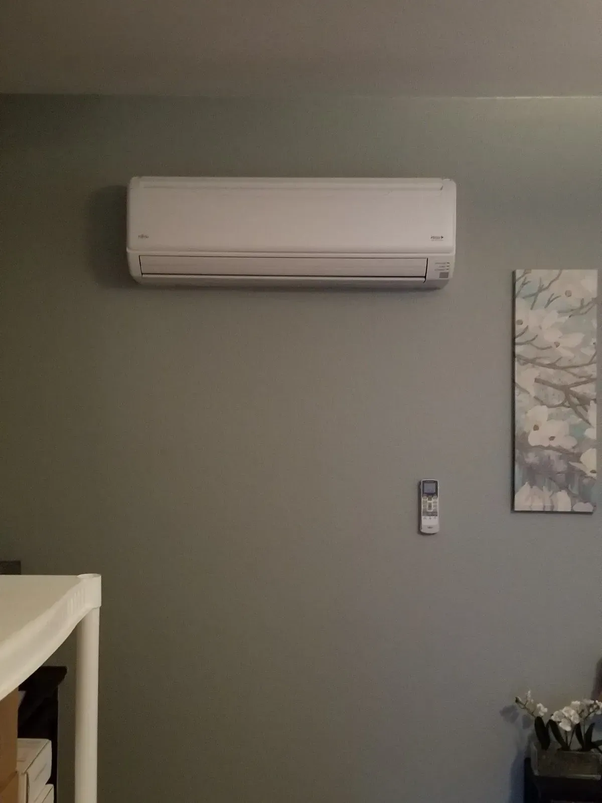 White wall-mounted air conditioning unit on a gray wall, below a remote control and art.