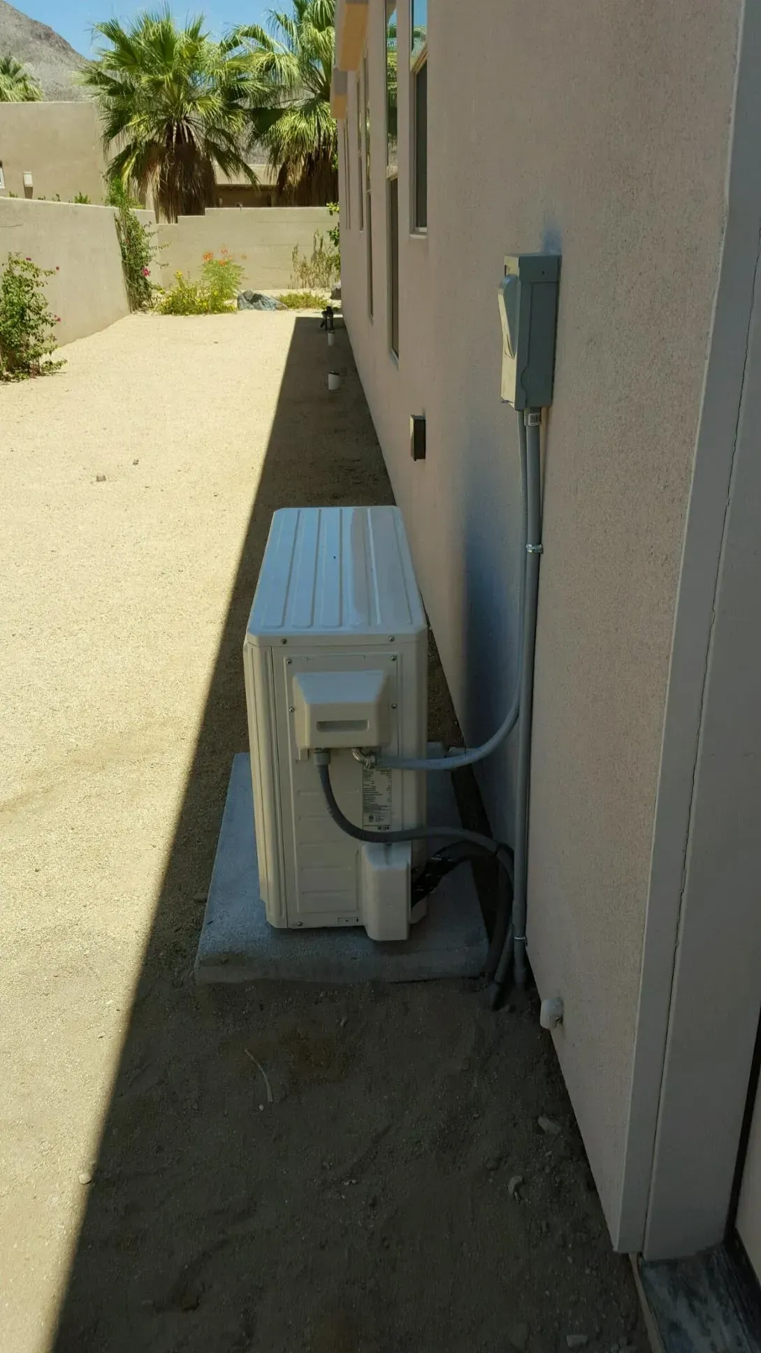 Air conditioning unit next to a light-colored wall and electrical box. Outdoor desert setting.