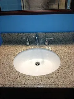 Oval bathroom sink with faucet and handles set in a speckled countertop against a blue wall.