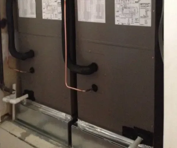 HVAC system with black insulated pipes and copper tubing against a grey wall.