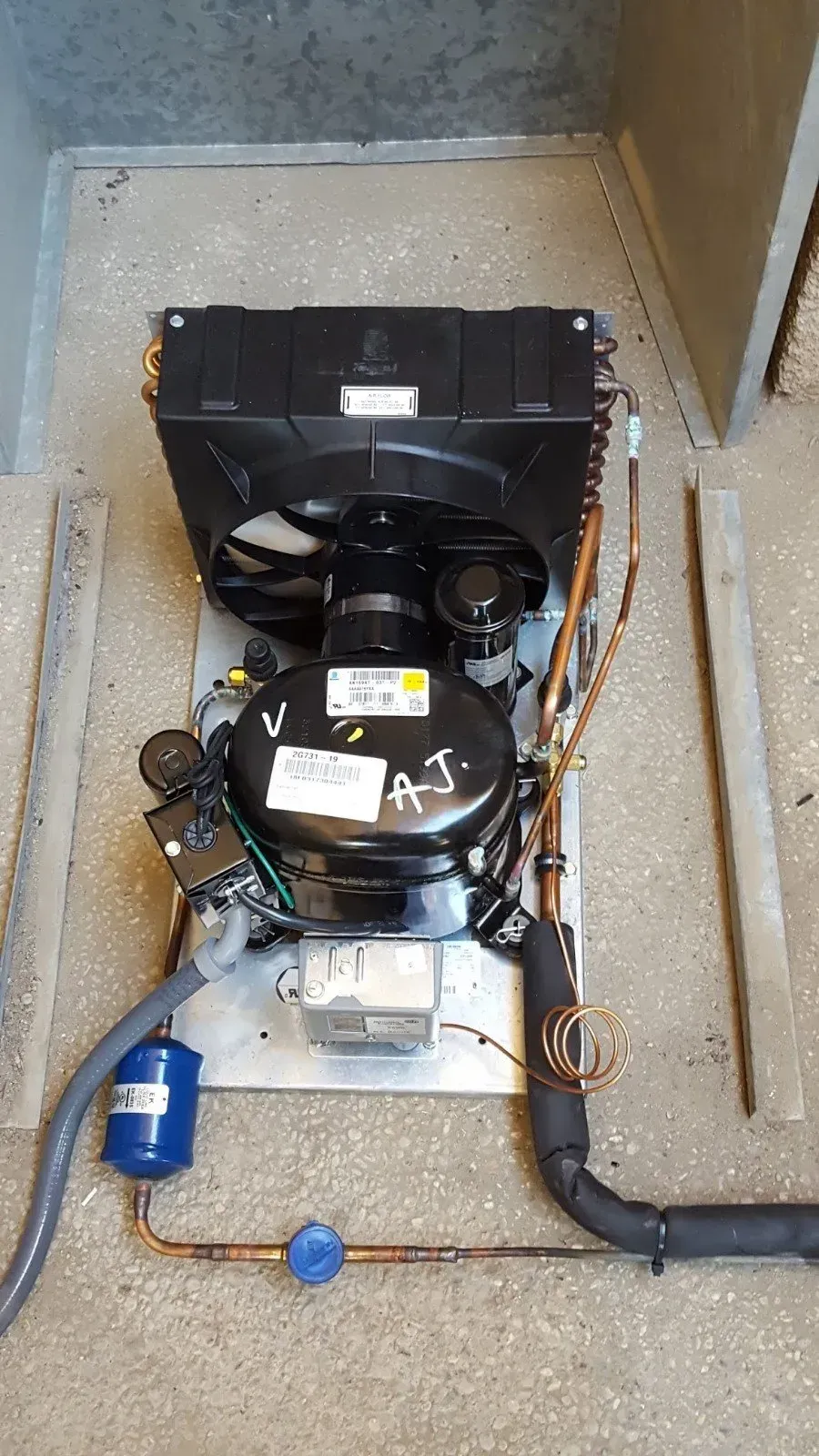 Refrigeration unit with black compressor, fan, and copper tubing on a metal base, in a gray setting.