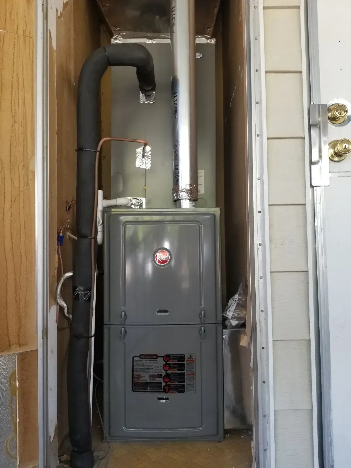 A Rheem furnace installed in a narrow alcove. The unit is gray with a silver exhaust pipe.