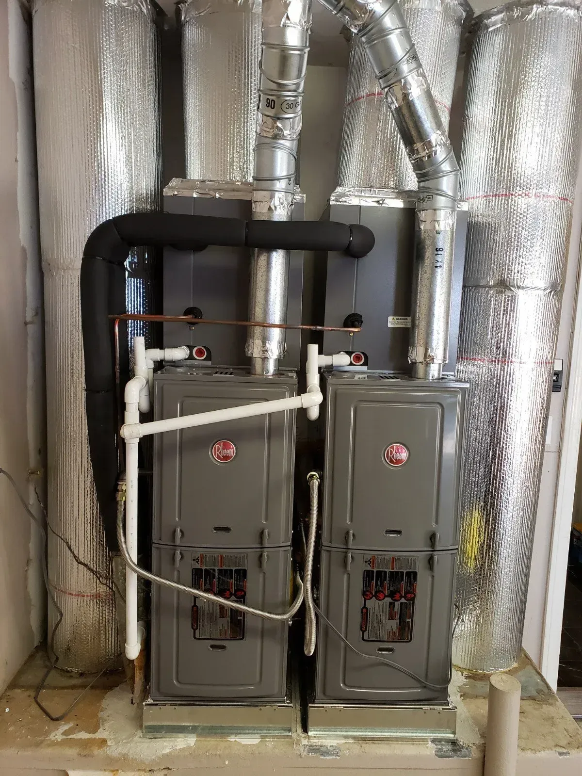 Two gray Rheem furnaces with ductwork in a utility room.