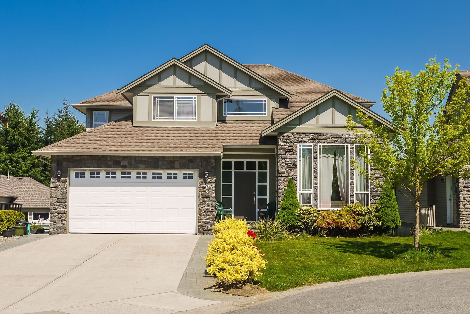 Two-story house with stone and tan siding, white garage door, and green lawn under a blue sky.
