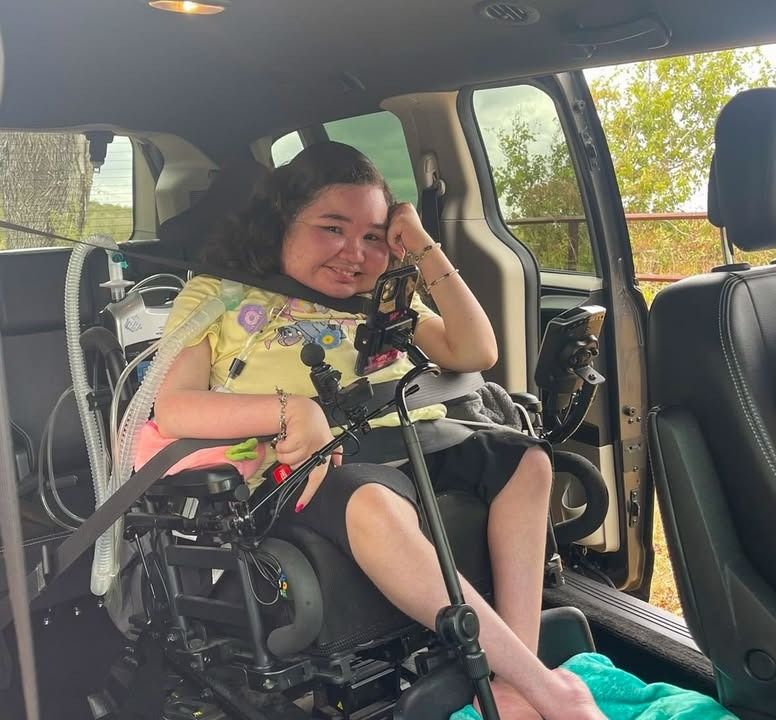 A smiling person in a wheelchair in a vehicle, wearing a yellow shirt.
