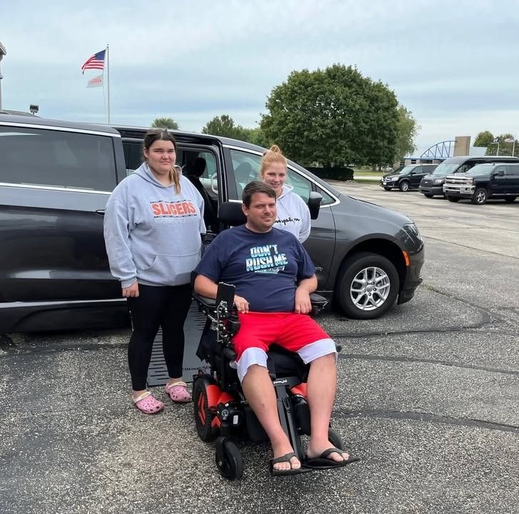 Man in wheelchair with two women by accessible van in parking lot.