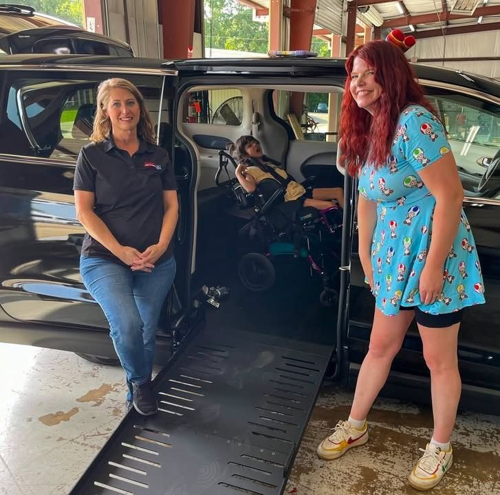 Two women and person in a wheelchair near a vehicle ramp. One woman wears a black shirt, the other a blue dress.