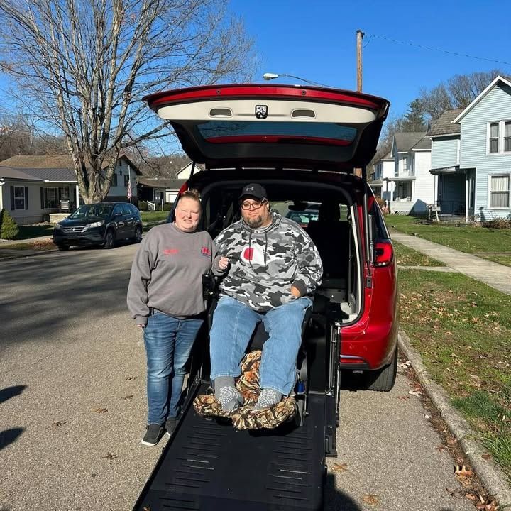 Woman and man in wheelchair by red van with ramp deployed on a residential street.