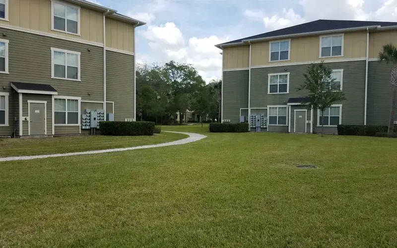 Two-story apartment buildings with green and tan siding, a grassy lawn, and a winding walkway.