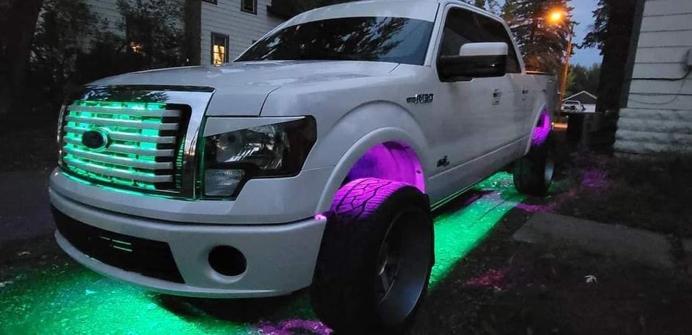A white truck with purple and green lights on it is parked in front of a house.