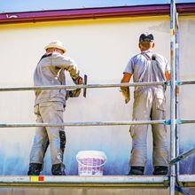 Two men are standing on a scaffolding painting a wall