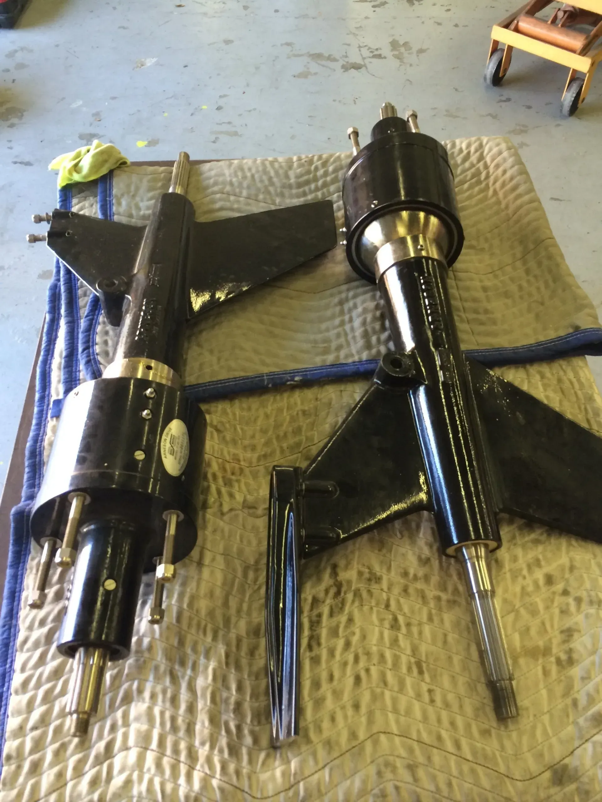Two black boat rudders with silver shafts and fins, on a blue and brown mat in a workshop.