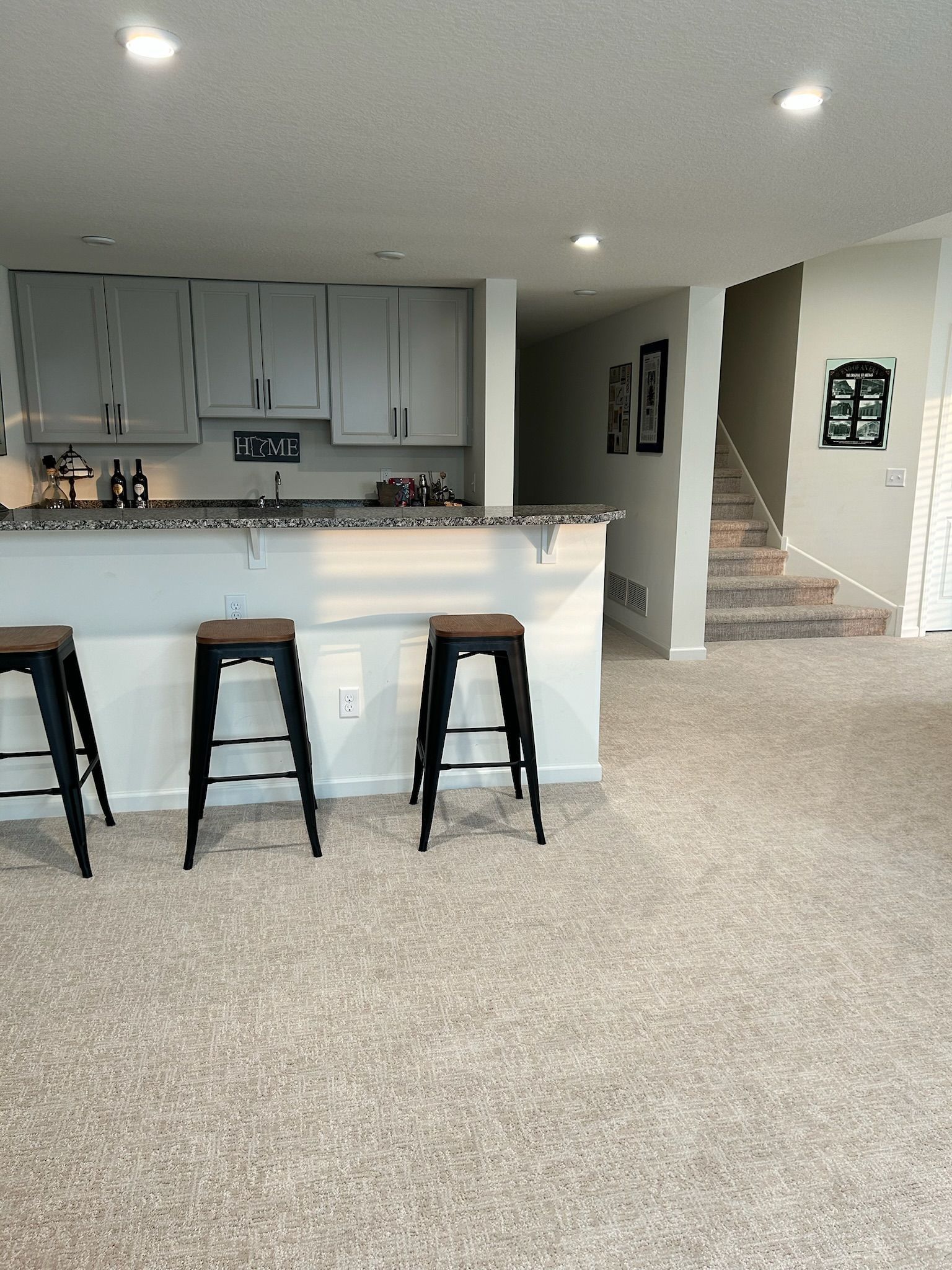 Carpeted living room with a kitchen island, three stools, and stairs leading up on the right.
