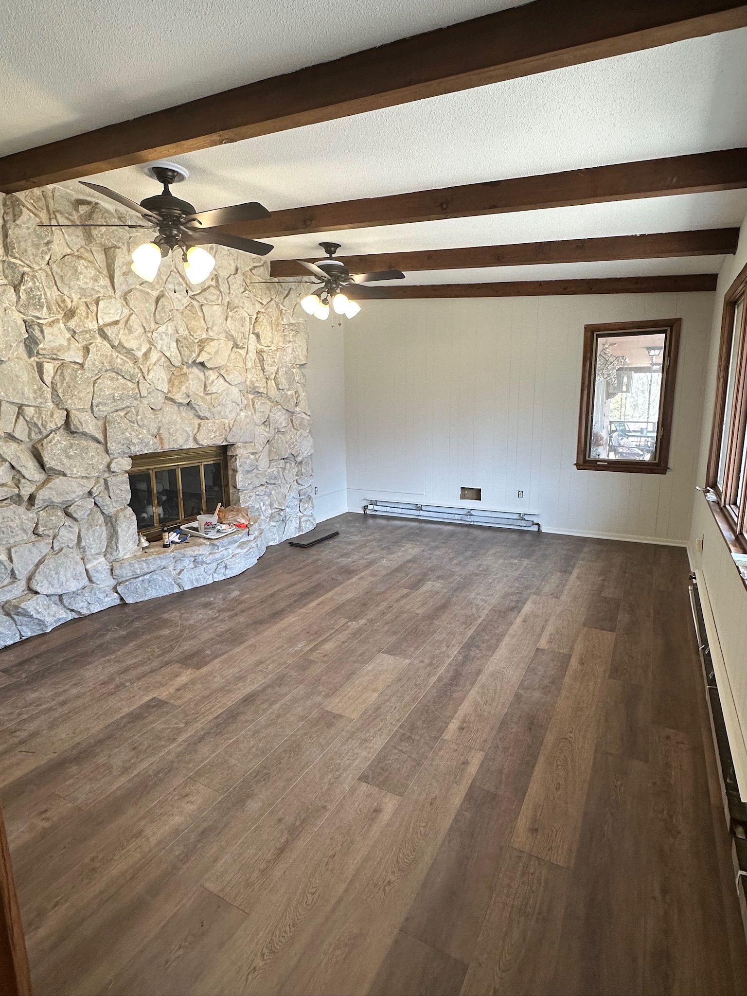 Empty living room with stone fireplace, wood beams, hardwood floor, and a window at the far end.