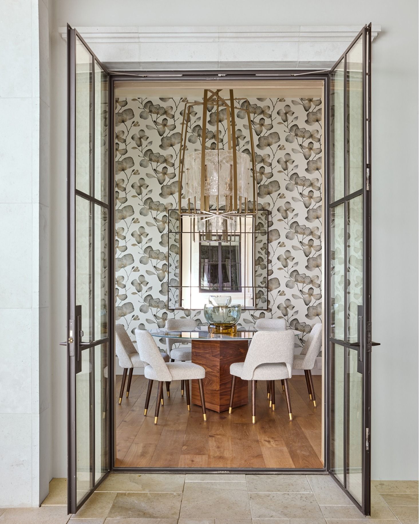 A dining room with a patterned wallpaper, modern chandelier, wooden table, and white chairs seen through glass doors.