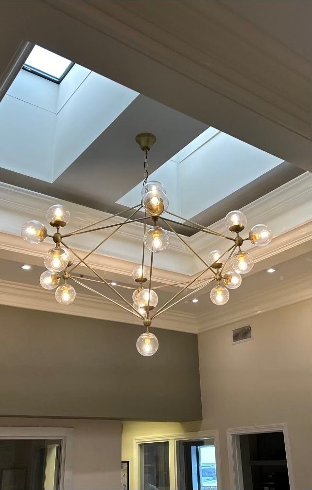 Geometric gold chandelier with glass globe lights hangs below a skylight in a room with white crown molding.