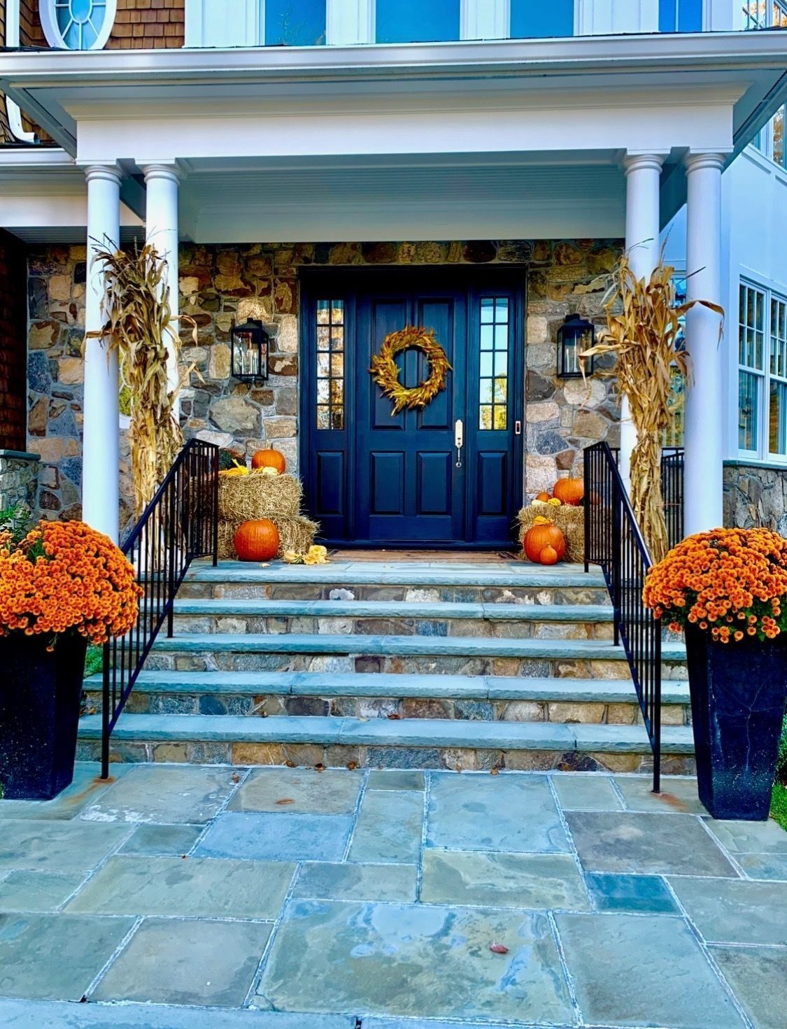 Stone front steps lead to a dark blue door decorated with fall decor: pumpkins, mums, and a wreath.