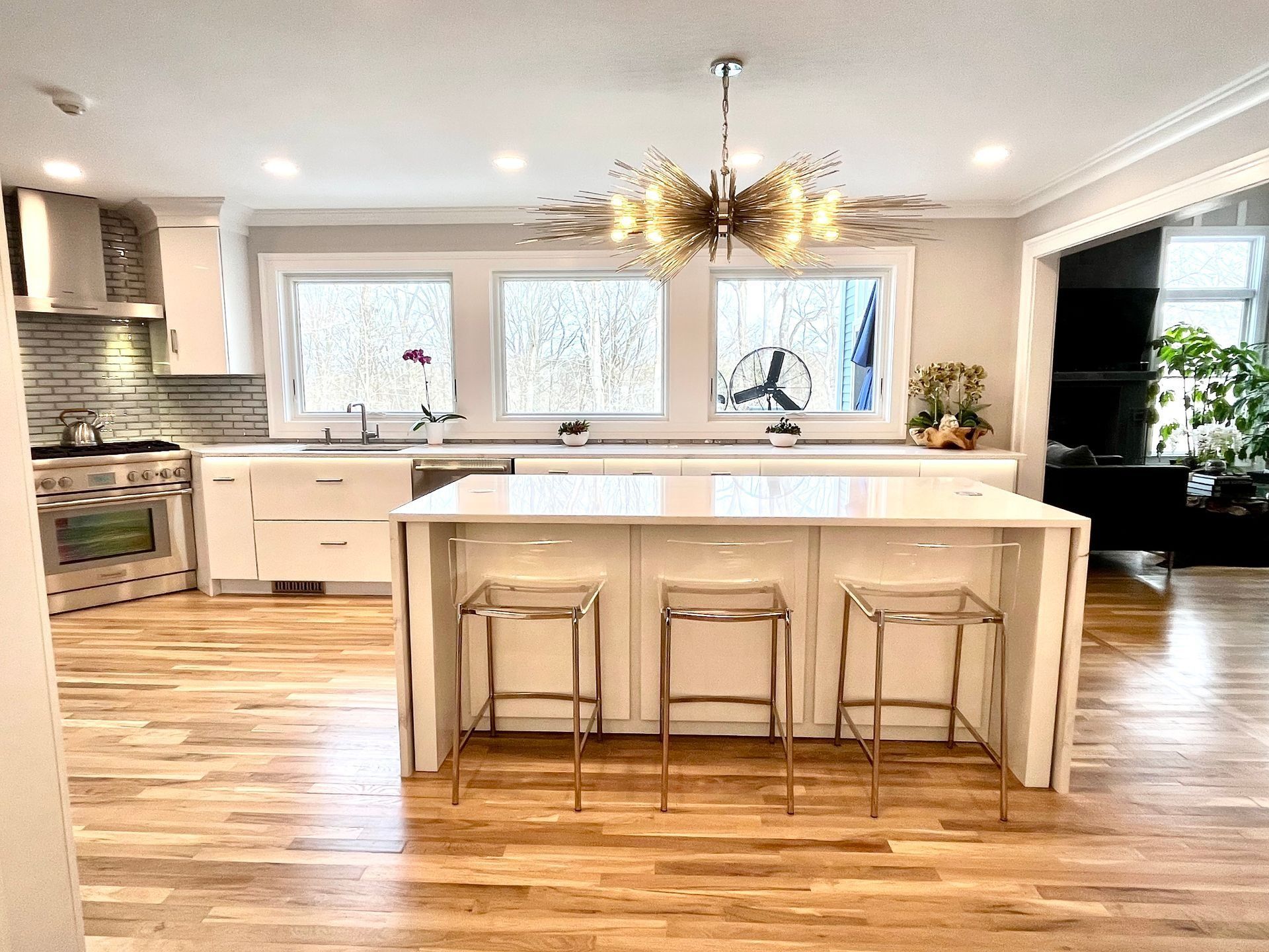 Modern kitchen with island, three bar stools, and chandelier. White cabinetry, wood floors, and windows.