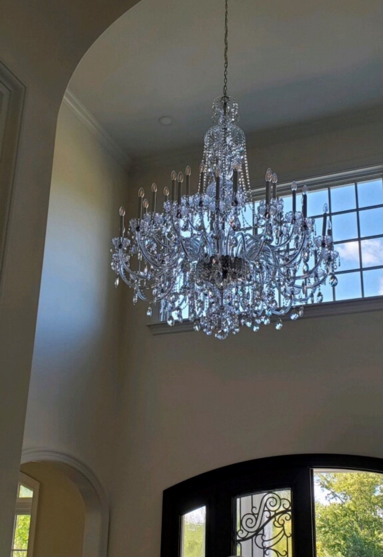 Crystal chandelier hanging in a large entryway with tall windows, reflecting sunlight.