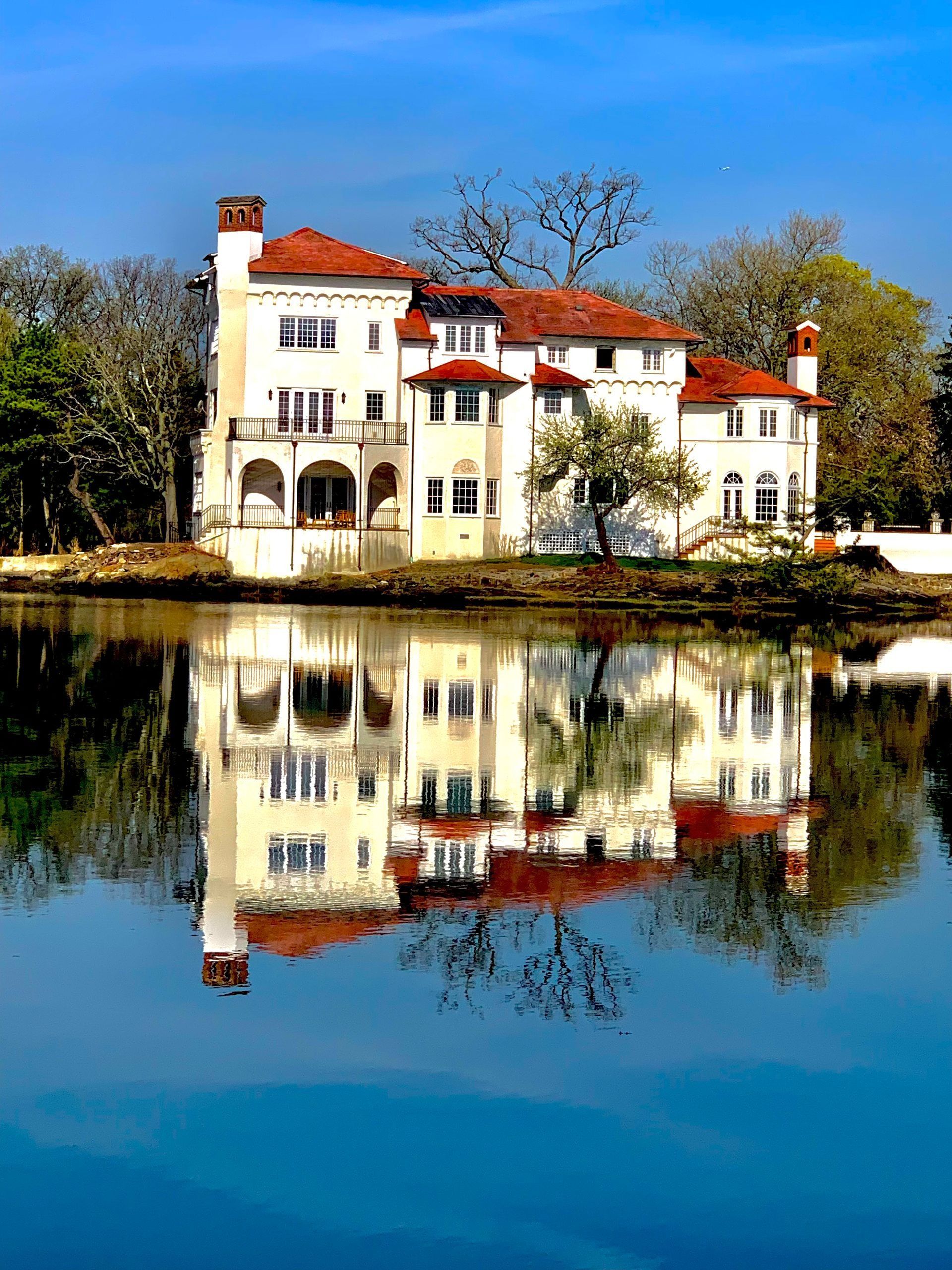 Elegant white mansion with red tile roof reflected in calm blue water.
