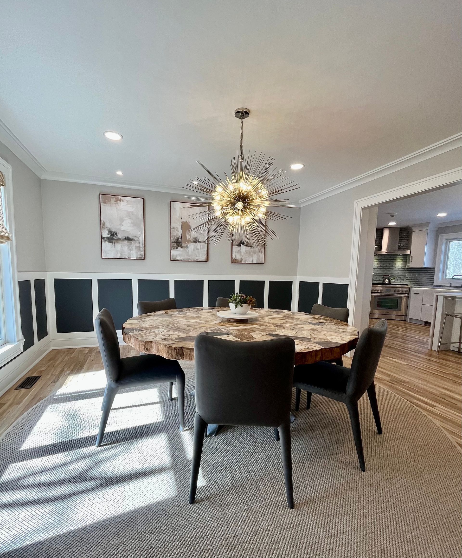 Dining room with round table, chairs, art, and modern chandelier. Gray and dark blue walls, light wood floor, and area rug.