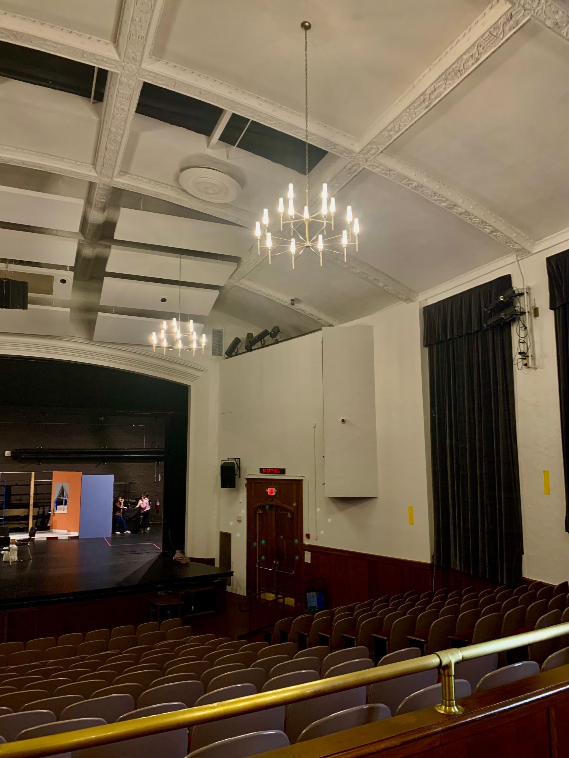 Auditorium interior with chandeliers, rows of seats, and stage with open black curtain.