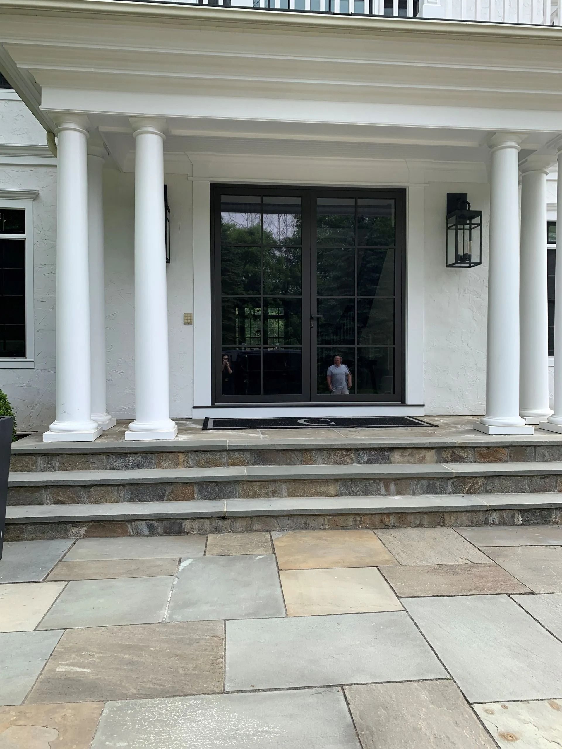 Exterior of a white house with a stone patio, steps, and dark double doors with a person visible inside.