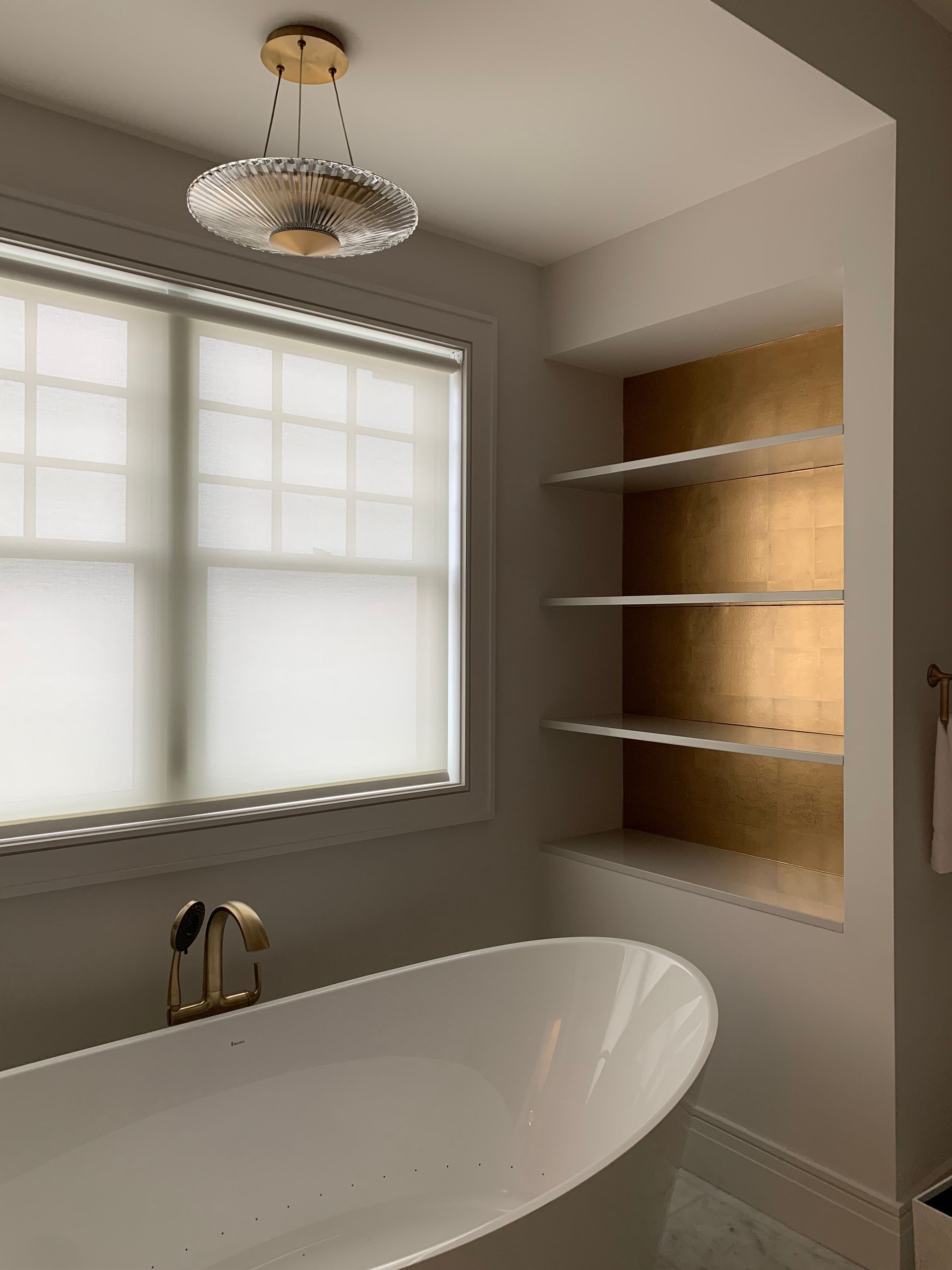 Bathroom with gold accents: white tub, shelves, gold faucet and light fixture, window with blinds.