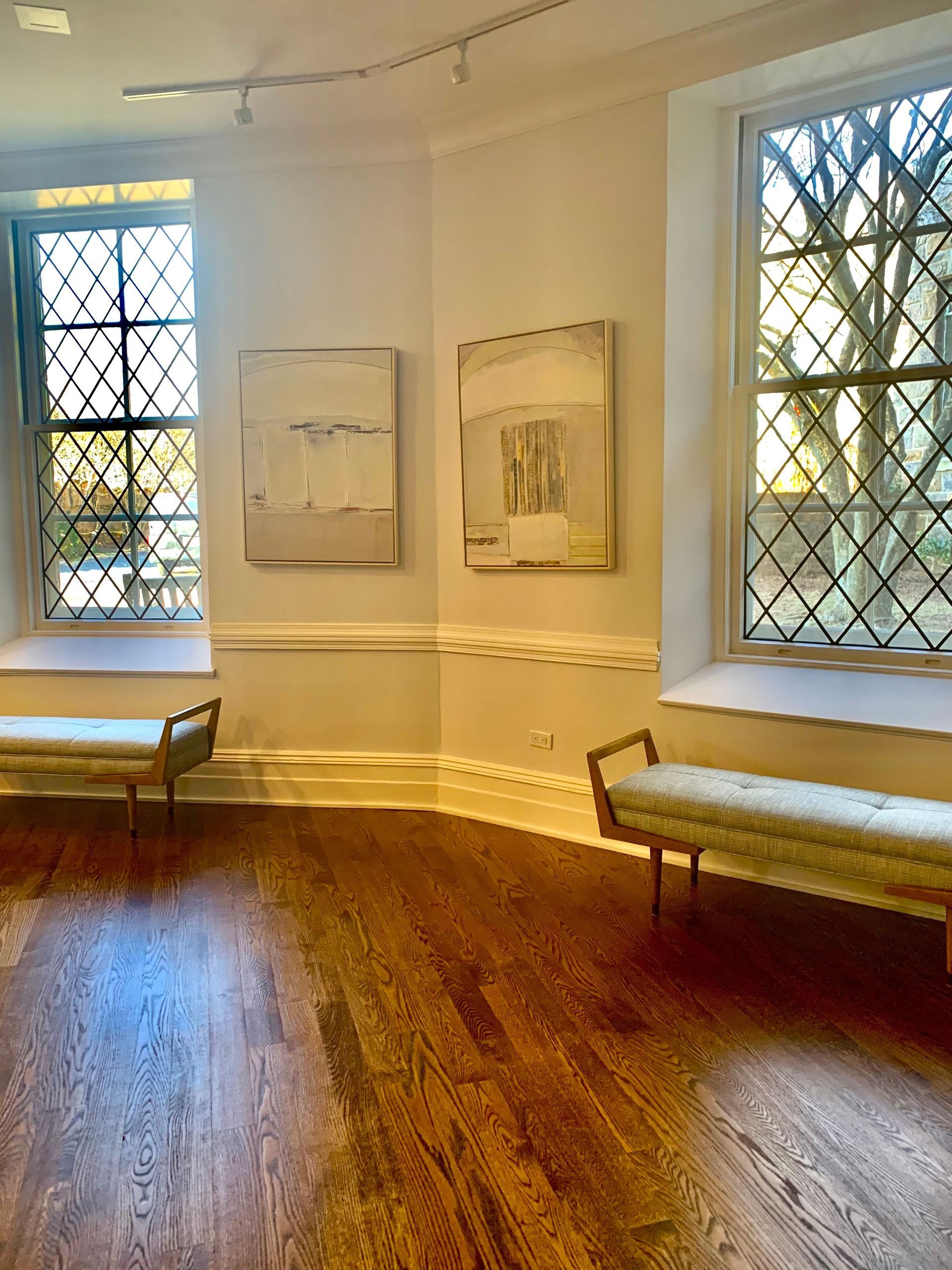Art gallery interior with abstract paintings, two windows, two benches, and hardwood floors.
