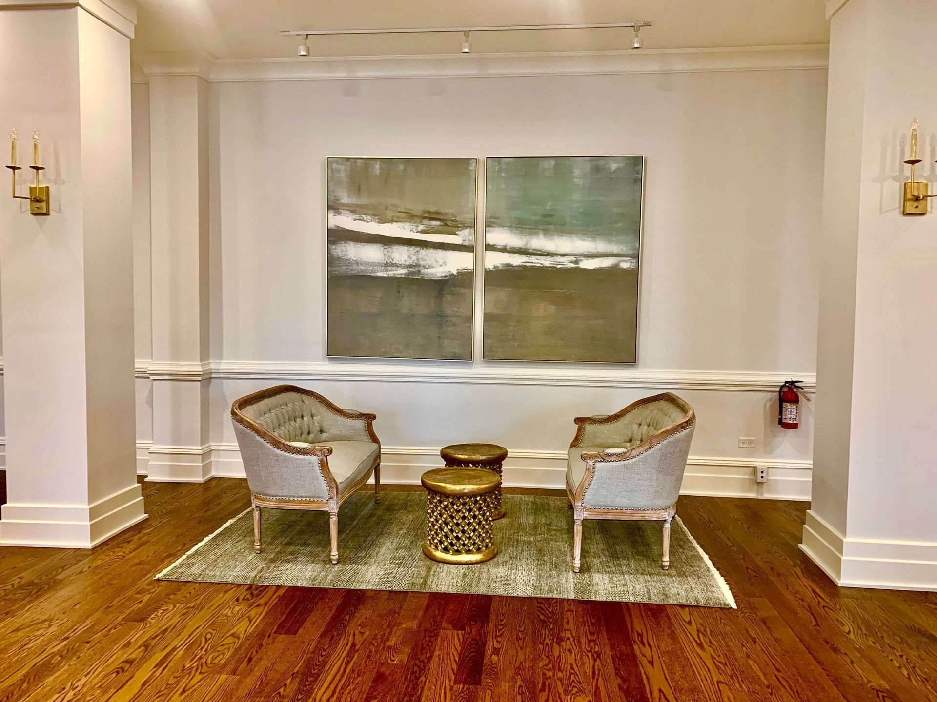 Two ornate chairs and two gold side tables on rug in front of abstract art on white wall with sconces, hardwood floor.