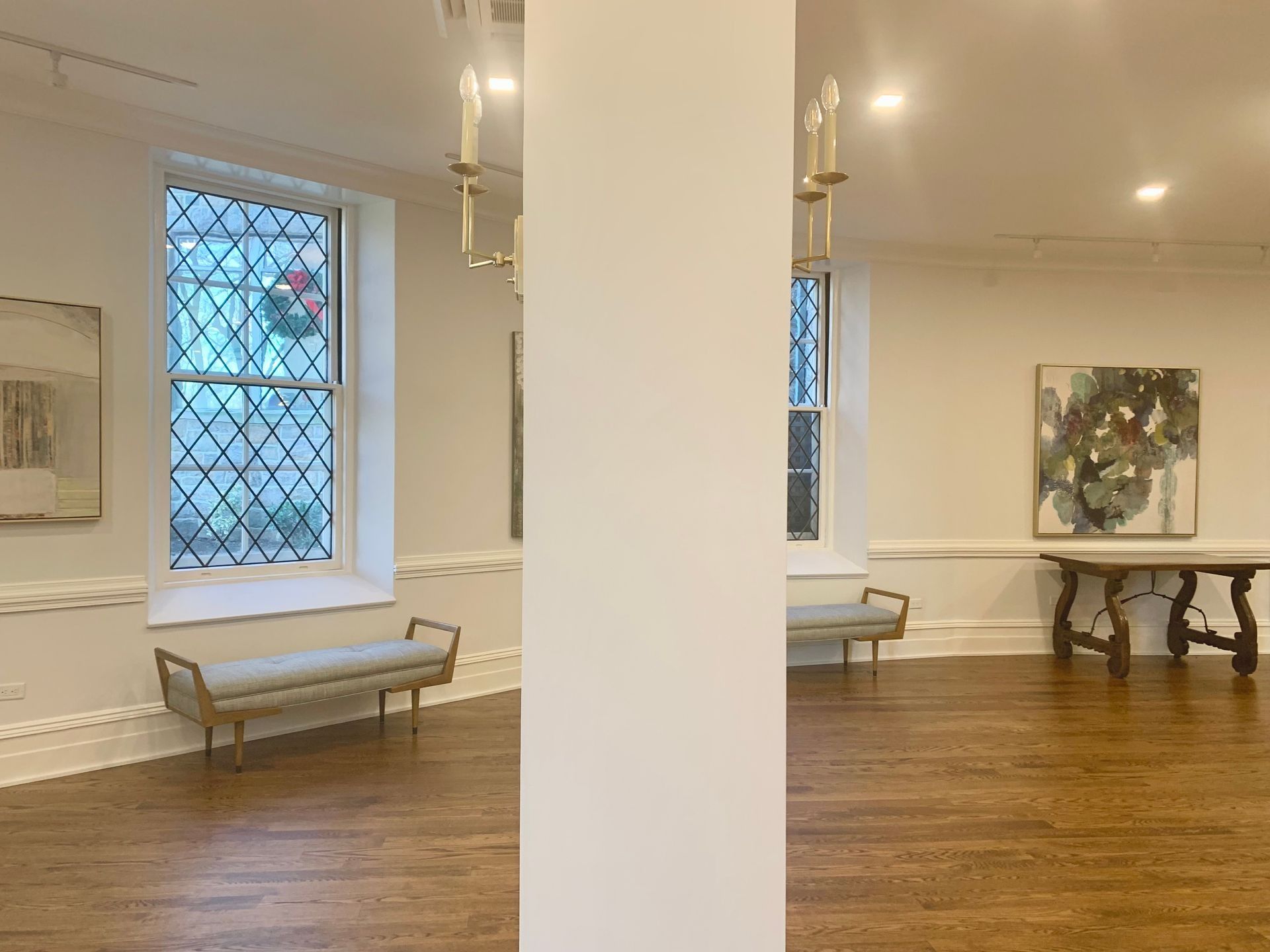 An art gallery with white walls, a wooden floor, and displayed paintings. Two benches sit by the windows.