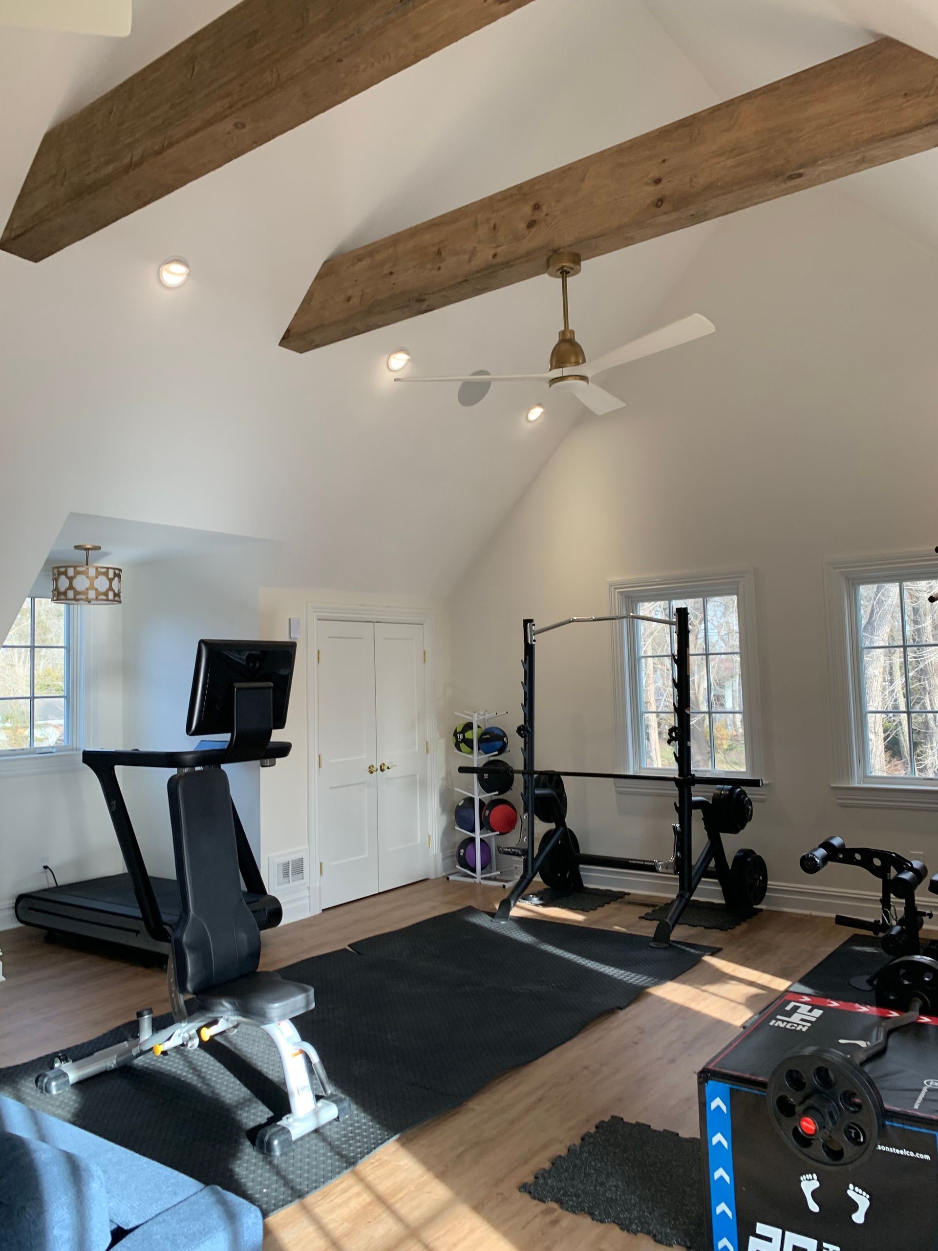 Home gym with treadmill, weight rack, dumbbells, and exercise equipment. Wooden beams on the ceiling, natural light.