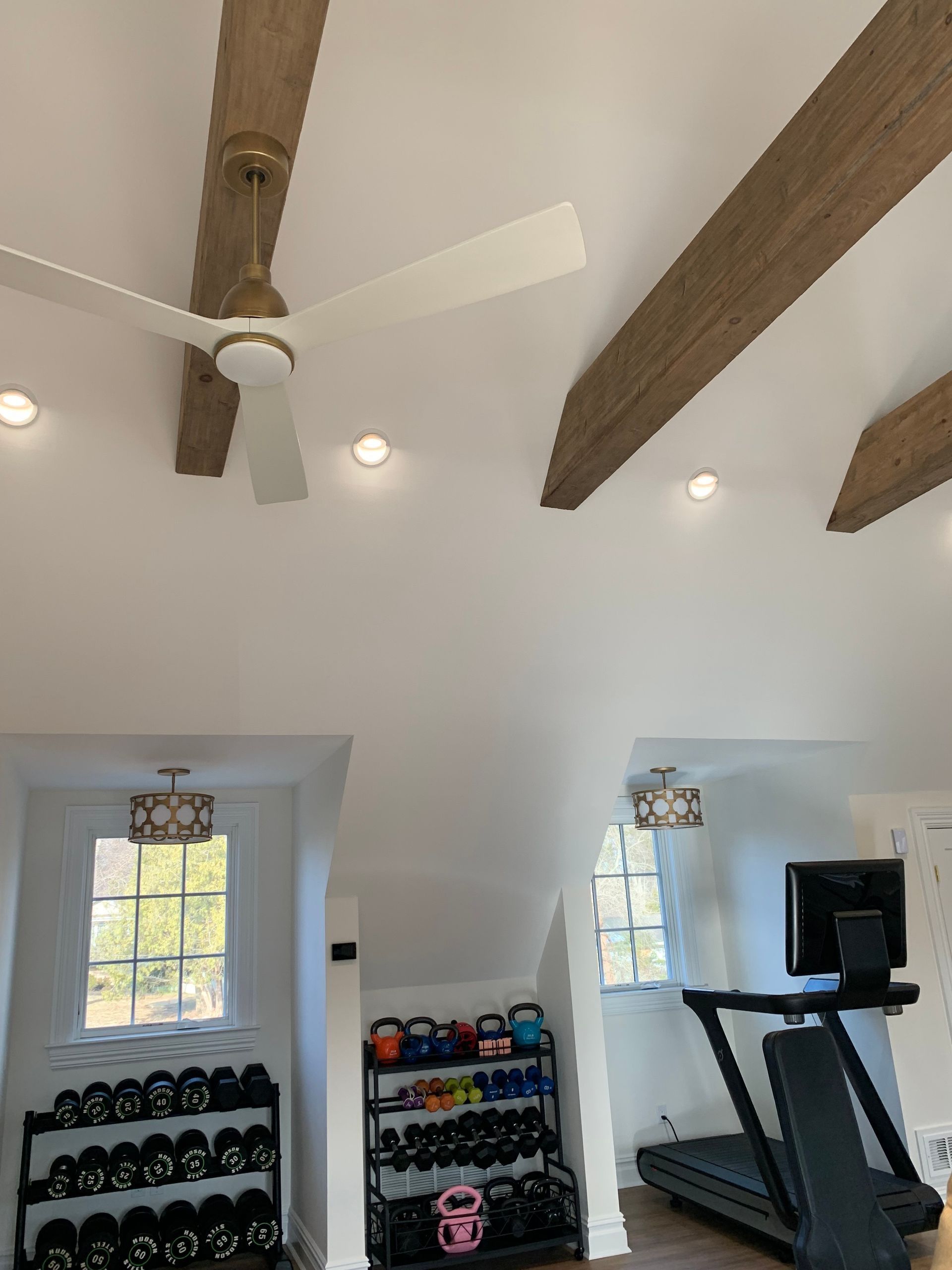 Home gym with weights, treadmill, and ceiling fan. White walls, wooden beams, windows, and recessed lights.