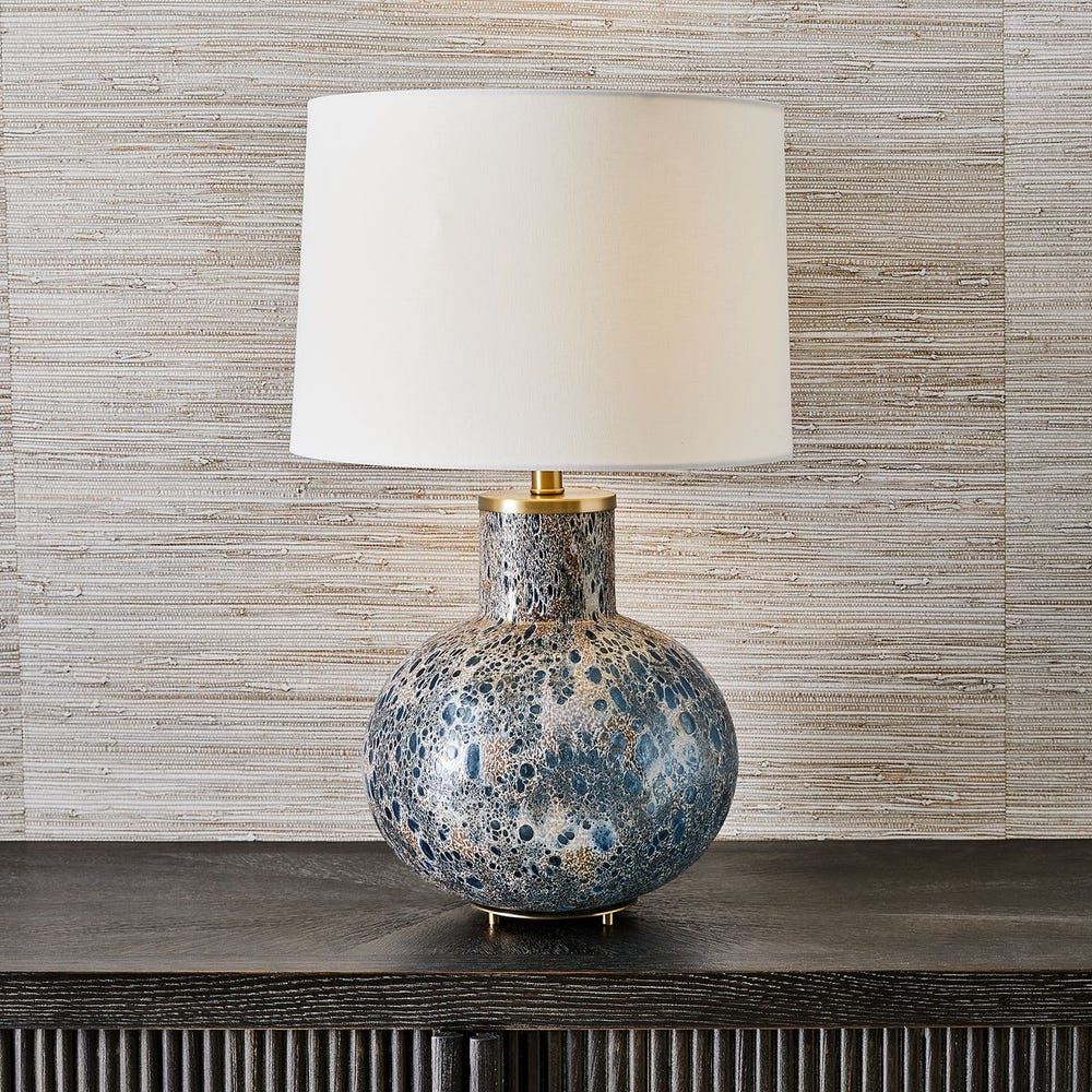 A table lamp with a blue, gold, and white speckled round base and a white drum shade, sitting on a dark wood surface.