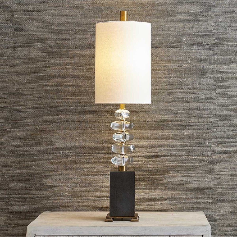 A tall table lamp with a dark rectangular base, stacked crystal accents on a gold stem, and a white cylindrical shade.