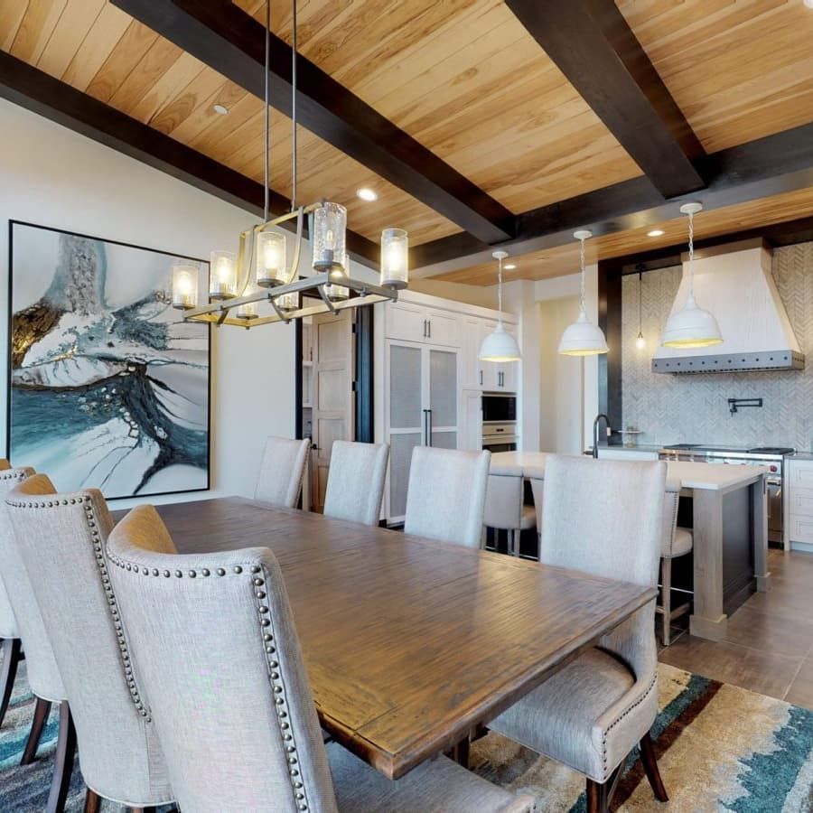 A dining area with a wood table and beige upholstered chairs beneath a lantern chandelier, opening into a modern kitchen.