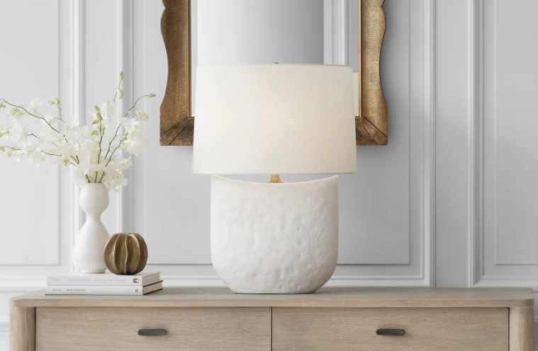 White table lamp on a light wood dresser with a vase of white flowers and a framed mirror behind it