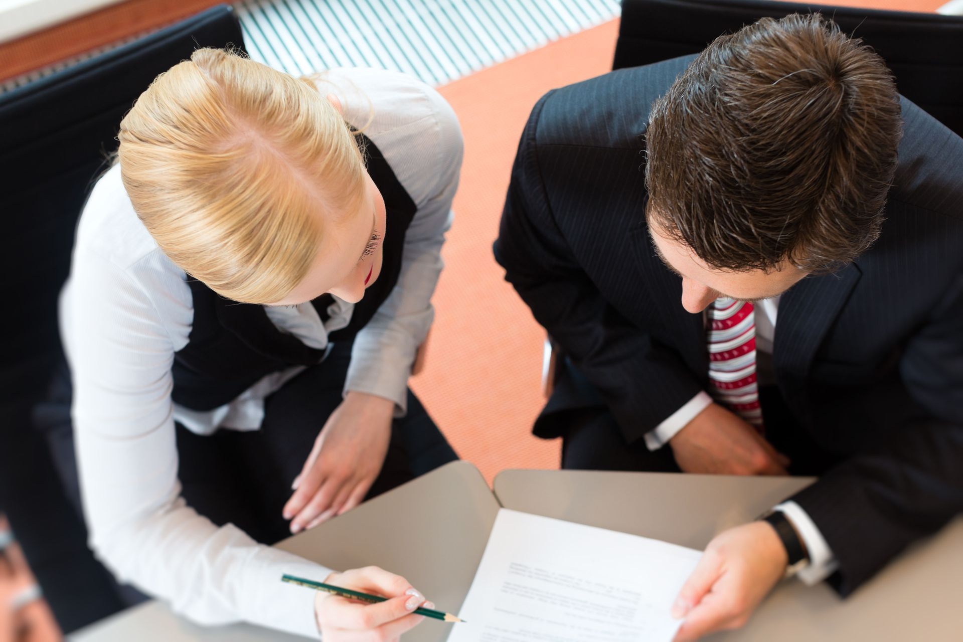 Two people looking at documents at a table. Woman points with pen. Man wears a suit.
